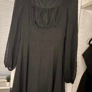 Svart långklänning med prickar - Säljer en svart långklänning med diskret prickigt mönster. Klänningen har transparenta långärmar med resår vid ärmslut och en lätt rynkad detalj vid halsen. Tyget är tunt och ger en luftig känsla, perfekt för dig som gillar stilrena och klassiska looks.