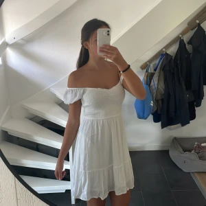 Vit kort klänning från Hollister i storlek S - Supersöt vit klänning från Hollister i storlek S. Den har smockad överdel, puffärmar som kan bäras offshoulder eller på axeln, och en volangkant nedtill. Klänningen är lätt och luftig, perfekt för varma dagar. Materialet känns som bomull och ger en skön känsla.