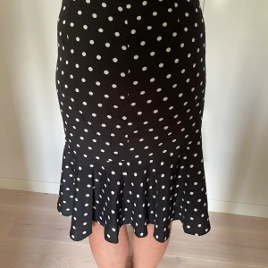 Svart prickig kjol med volang - Supersnygg svart kjol med vita prickar och volang nertill. Kjolen har en figurnära passform upptill och släpper ut i en volang som ger en lekfull vibe. Perfekt för dig som gillar retrostil och vill sticka ut lite extra.