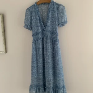 Blå leopardmönstrad klänning  längre modell  - Säljer en blå kortklänning med vitt leopardmönster och omlott-v-ringning. Klänningen har volangdetaljer längs halsringning, midja och nederkant samt korta puffärmar med volang. Perfekt för sommarens alla tillfällen!