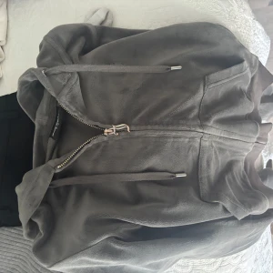 Grå fleecejacka med huva och dragkedja - Mysig grå fleecejacka från Juicy Couture med huva, dragkedja och snörning. Jackan har stora fickor framtill och ribbade muddar. Perfekt för chill dagar eller när du vill vara extra bekväm.