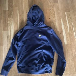 Marinblå hoodie från Lyle & Scott - Snygg marinblå hoodie från Lyle & Scott med klassisk gul fågel-logga broderad på bröstet. Tröjan har huva, känguruficka och långa ärmar. Perfekt för chill dagar och enkel att matcha med jeans eller joggers. Mjuk bomullskvalitet som känns skön mot huden.
