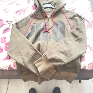 Olivgrön hoodie med röd snörning Emilio - Olivgrön hoodie från Emilio med stor svart print 'PILOT' på framsidan. Röd snörning och sömmar ger en cool kontrast. Dragkedja framtill och huva för extra chill vibe. Mjuk insida och ribbade muddar vid ärmslut och nederkant. Perfekt för dig som gillar streetwear.