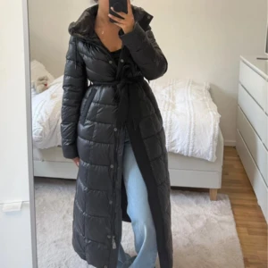 Lång svart dunjacka med bälte - Snygg lång svart dunjacka med quiltad design och bälte i midjan. Jackan har hög krage, knappar och dragkedja framtill. Perfekt för kalla dagar och ger en stilren siluett. Passar dig som vill ha både värme och trendig look.