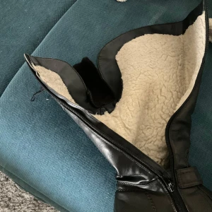 Svarta fodrade stövlar med spänne - Svarta stövlar i skinn med mysigt vitt foder och dragkedja på sidan. Snygga detaljer med spänne och dekorativa sömmar. Grov sula och låg klack gör dem perfekta för kyliga dagar.