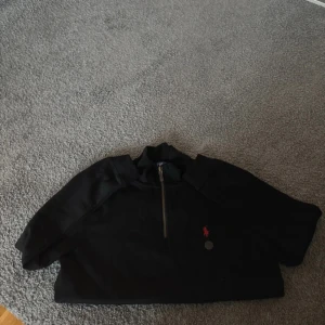 Svart half zip hoodie från Polo Ralph Lauren - Svart hoodie från Polo Ralph Lauren med röd broderad logga på bröstet. Tröjan har half zip framtill och är tillverkad i mjuk bomull. Långärmad och med klassisk krage, perfekt för chill dagar. Snygg och enkel att matcha med jeans eller joggers.