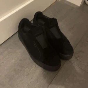 Axel arigato skor  - Stilrena svarta slip-on sneakers med ovandel i mocka och elastiska paneler för enkel påtagning. Skorna har en platt sula och rund tå, vilket ger en clean och modern look. Perfekta för dig som gillar minimalistisk stil.
