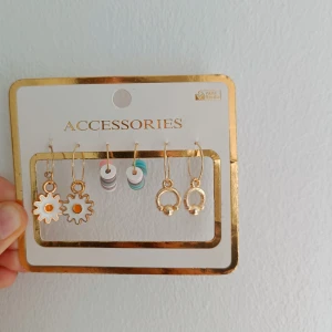 3-pack örhängen i guld och färg - Säljer ett 3-pack örhängen från Accessories. Ett par med blommor i guld och vitt med orange detalj, ett par med färgglada plattor i vitt, rosa, grått och turkost, samt ett par små guldörhängen med rund form och pärldetalj. Perfekt för att mixa och matcha din stil.