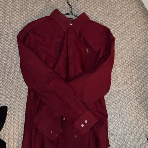 Vinröd skjorta från Ralph Lauren - Snygg vinröd skjorta från Ralph Lauren med klassisk krage och broderad logga på bröstet. Skjortan har långa ärmar och knappar framtill. Perfekt för dig som vill ha en stilren och tidlös look.