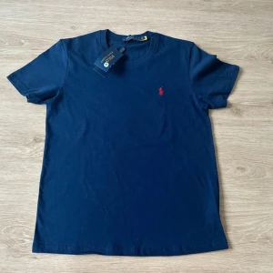Ralph Lauren T-Shirt - Helt oanvänd! - Helt oanvänd Ralph Lauren T-Shirt säljes. Tröjan är blå med med den klassiska logotypen broderad i rött på vänster bröst! | Skick: 10 / 10 | Nypris: ~ 1100 | Vårt pris: 499 | Storlek: M | Tveka inte att höra av er med funderingar!