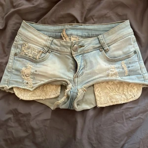 Lågmidjade  jeansshorts med spetsdetaljer - Supersnygga ljusblå jeansshorts med slitna detaljer, pärlor och spetsinfällningar på sidorna. Jag säljer dom pga ingen användning! 