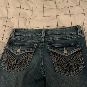 Blå jeans med broderade bakfickor Gina Tricot - Snygga blå jeans från Gina Tricot med coola broderade detaljer på bakfickorna och kontrastsömmar. Klassisk femficksmodell med knapp och dragkedja framtill. Perfekta för dig som vill sticka ut lite extra med dina jeans.