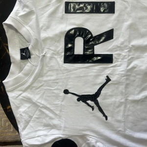 Jordan x Paris vit t-shirt - Snygg vit t-shirt från Jordan med PARIS-tryck i svart och den ikoniska Jumpman-loggan broderad på bröstet. T-shirten har rund hals och korta ärmar, tillverkad i mjuk bomull. Perfekt för dig som gillar sportig streetstyle och vill sticka ut.