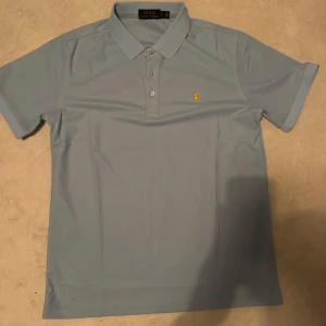 Ljusblå Polo Ralph Lauren t-shirt - Snygg ljusblå t-shirt från Polo Ralph Lauren med klassisk pikékrage och korta ärmar. T-shirten har två knappar framtill och den ikoniska gula Polo-loggan broderad på bröstet. Perfekt för dig som gillar stilren och enkel design.