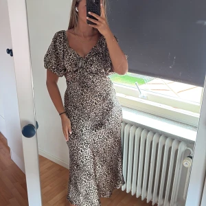 Leopardmönstrad långklänning med puffärm - Snygg långklänning med leopardmönster och puffiga korta ärmar. Klänningen har v-ringning med knyt framtill och är figurnära upptill med lätt utsvängd kjol. Materialet är glansigt och känns lyxigt, perfekt för dig som vill sticka ut. Helt helt ny med prislapp kvar returneringstiden var över när jag kom hem från resa.