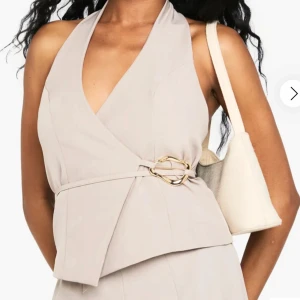 Beige tailored omlottopp från Boohoo - Snygg beige omlottopp från Boohoo med halterneck och öppen rygg. Toppet har en dekorativ guldspänne i midjan och är ärmlös. Perfekt för dig som gillar stilrena och trendiga detaljer. Helt helt ny och oanvänd med prislapp kvar min returneringstid var över när jag väl var hemma från semester tyvärr.
