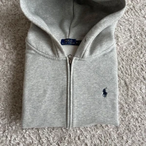 Grå hoodie från Polo Ralph Lauren - Snygg ljusgrå hoodie från Polo Ralph Lauren med dragkedja och klassisk broderad logga i mörkblått på bröstet. Tröjan har huva, känguruficka och är tillverkad i mjuk bomullsmix. Perfekt för chill dagar eller när du vill ha en clean look.