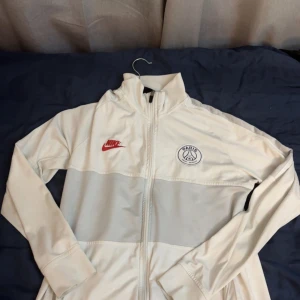 Vit Nike PSG  med dragkedja - Snygg vit vindjacka från Nike med PSG-logga på bröstet och röd Nike-swoosh. Koftan har hel dragkedja framtill, hög krage och långa ärmar. Perfekt för dig som gillar sportig stil och vill representera Paris Saint-Germain.