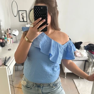 Blå topp med volang från Lindex - Supersöt ljusblå topp från Lindex med en axel och volangdetalj. Tunt, strukturerat tyg som känns luftigt och perfekt för varma dagar. Sitter snyggt med omlottkänsla. Perfekt att styla med jeans eller kjol.