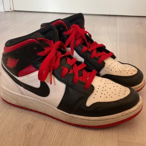 Nike Air Jordan 1 svart/vit/röd - Säljer ett par Nike Air Jordan 1 sneakers i klassisk svart, vit och röd färg. Skorna har röda snören, svart swoosh och Air Jordan-logga på sidan. Tillverkade i skinn med perforerad tå och vadderad häl för extra komfort. Perfekta för dig som gillar streetstyle.