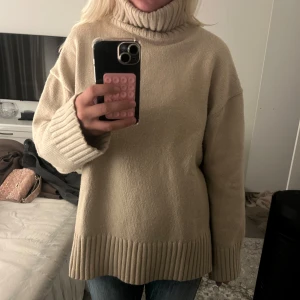 Beige polotröja i stickad ull - Supermysig beige polotröja i stickad ull med oversized passform. Tröjan har breda ribbade muddar vid ärmslut och nederkant samt hög polokrage. Perfekt för kalla dagar och ger en chill vibe till din outfit.