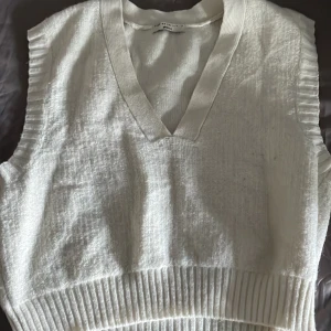 Vit stickad väst från Gina Tricot - Säljer en vit stickad väst från Gina Tricot i storlek S. Västen har v-ringning, bred ribbad kant vid ärmhål och nederkant samt en mjuk, lite fluffig struktur. Perfekt att styla över en skjorta eller t-shirt för en trendig look.