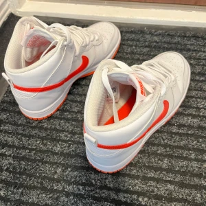 Nike Dunk Mid vit/röd sneakers - Säljer ett par nike dunk mid sneakers i vitt med orange swoosh och detaljer. Använd väldigt fåtal gånger, som ny! 