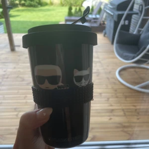 Karl Lagerfeld  -  en svart take-away mugg med illustrationer av en person med solglasögon och en katt med solglasögon. Muggen har en gummiring för grepp och är märkt med 'Karl Lagerfeld'.