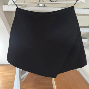 Svart omlottkjol från Zara - Stilren svart kortkjol med inbyggda shorts från Zara i omlottmodell. Kjolen har minimalistisk design med två fuskfickor bak och stängs med dragkedja i sidan. Perfekt för dig som gillar en clean och modern look. Prislapp kvar, nypris 329kr