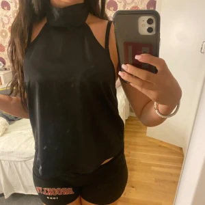 Svart polotopp med knyt i ryggen - Säljer en svart polotopp med halterneck och snygg knytning i ryggen. Toppen är ärmlös och har en öppen rygg som ger en cool vibe. Materialet känns mjukt och har ett lätt glansigt utseende, perfekt för att sticka ut lite extra.