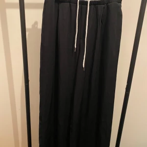 Svarta wide byxor från Mussera - Svarta byxor från Mussera med bred passform och resår i midjan. Byxorna har vita snören för justering och två bakfickor. Materialet känns lätt och flowy, perfekt för en chill och avslappnad look.