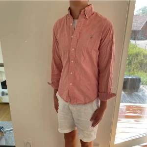 Rosa skjorta från Ralph Lauren - Snygg rosa skjorta från Ralph Lauren med klassisk button down-krage och broderad logga på bröstet. Skjortan har lång ärm, knappar framtill och är tillverkad i mjuk bomull. Perfekt för en chill och stilren look.