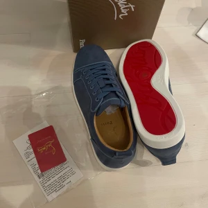 Blå sneakers från Christian Louboutin - Snygga blå sneakers från Christian Louboutin i mocka med klassisk röd sula och vita kanter. Skorna har rund tå, snörning och exklusiv känsla. Perfekta för dig som vill sticka ut med stilrena detaljer och lyxig design.