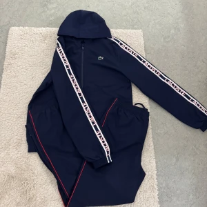 Lacoste dress - Snygg marinblå vindjacka från Lacoste med huva och dragkedja. Jackan har vita Lacoste-loggor längs ärmarna och diskret krokodillogo på bröstet.                                                        Storlek M