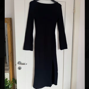 Svart långärmad ribbad klänning - Snygg svart långklänning med ribbad struktur och långärmade ärmar. Klänningen har en slits på ena sidan och en djupare rygg, vilket ger en cool och stilren look. Perfekt för dig som gillar minimalistisk stil med en twist.