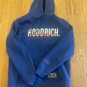 Blå hoodie från Hoodrich med tryck - Säljer en blå hoodie från Hoodrich med stor vit och röd logga på bröstet och ryggen. Hoodien har huva med snörning, känguruficka och coolt streetwear-märke framtill. Röd kontrastsöm vid fickan och tryck på ärmen ger extra stilpoäng.