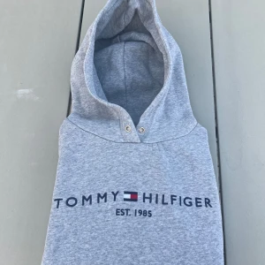 Grå hoodie Tommy Hilfiger M - Snygg grå hoodie från Tommy Hilfiger i storlek M. Tröjan har huva med metallöglor och klassisk logga tryckt framtill. Mjuk insida och skön passform, perfekt för chill eller häng med kompisar. Ikoniska Hilfiger-detaljer vid halsen.