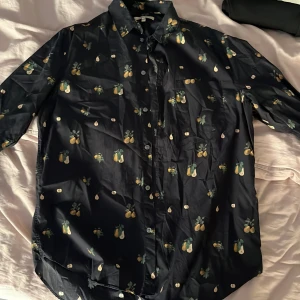 Mörkblå skjorta med päronmönster Whyred - Snygg mörkblå skjorta från Whyred med unikt päronmönster i grönt och gult. Skjortan är tillverkad i 100% bomull och har klassisk krage, knäppning framtill och långa ärmar. Perfekt för dig som vill sticka ut med en cool printad skjorta.