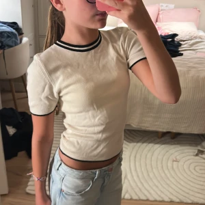 Vit ribbad topp från Gina Tricot XS - Säljer en vit ribbad topp från Gina Tricot i storlek XS. T-shirten har svarta ränder vid halsringning, ärmslut och nederkant för en sportig vibe. Modellen är croppad och har korta ärmar. Perfekt till jeans eller kjol för en clean look.