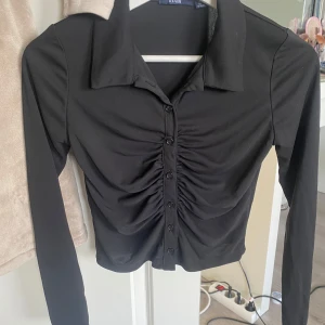 Svart långärmad blus från KIABI - Snygg svart blus från KIABI i storlek XS. Toppen har långa ärmar, klassisk krage och knappar framtill. Framsidan är rynkad för en cool effekt och modellen är croppad. Perfekt för dig som gillar stilrena och trendiga plagg.