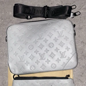 axelväska i silver - Snygg axelväska silverfärgat äkta skinn med klassiskt monogrammönster. Väskan har en bred svart justerbar axelrem och dragkedja upptill. Perfekt för dig som vill ha en stilren och lyxig accessoar.