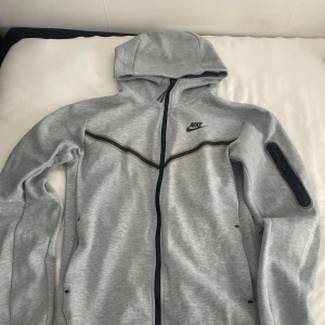 nike tech fleece - Snygg grå hoodiejacka från Nike med hel dragkedja och svart logga på bröstet. Jackan har huva, svarta detaljer längs bröstet och en praktisk ficka med dragkedja på ärmen. Perfekt för chill eller träning. Helt nya nästan Storlek S