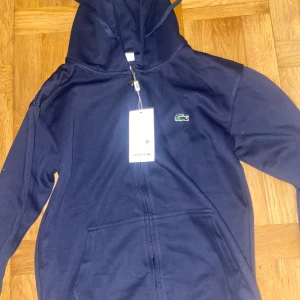 Marinblå hoodie från Lacoste M - Snygg marinblå hoodie från Lacoste med dragkedja och klassisk krokodillogga på bröstet. Tröjan har huva med snörning, känguruficka och regular fit. Perfekt för dig som gillar stilren och sportig look.
