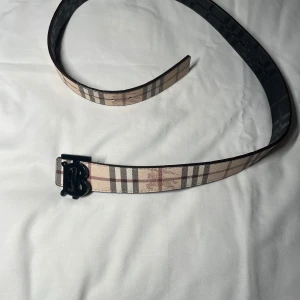 Burberry bälte 110cm - Snyggt Burberry-bälte i klassiskt rutigt mönster med beige, svart och röda toner. Bältet har ett stort svart TB-monogram som spänne och är tillverkat i skinn. Insidan är svart med präglat Burberry-mönster och logga. Perfekt accessoar för att lyfta din outfit. Finns två stycken 
