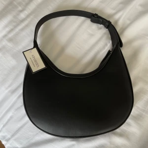 Svart axelväska från Ullared  - Stilren svart axelväska från Ullared med justerbart axelband och dragkedja upptill. Väskan har en mjukt rundad form och är gjord i slätt syntetmaterial. Perfekt storlek för det viktigaste och har ett innerfack med dragkedja och även två små fack till. Aldrig använd och nyligen köpt!