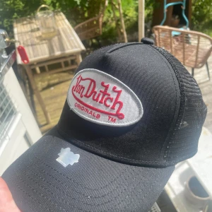 Svart Von Dutch truckerkeps - Säljer en svart truckerkeps från Von Dutch med klassisk mesh på baksidan och justerbar snapback. Framtill finns en stor broderad logga i vitt och rött. Insidan har coolt tryck med Von Dutch-motiv. Perfekt för dig som vill ha retrostil och streetvibe.