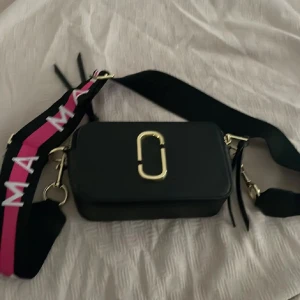 Svart axelväska från Marc Jacobs - Snygg svart axelväska från Marc Jacobs med guldiga metalldetaljer och dragkedja. Väskan har en bred axelrem med rosa och vita detaljer samt Marc Jacobs-logga. Tillverkad i skinn med stilren, boxig form och praktiskt fack på baksidan.