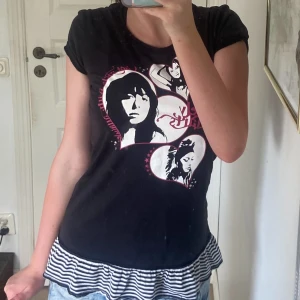 Svart t-shirt med hjärtmotiv och volang - Cool svart t-shirt från Wild Venus med tryck av ansikten i vita och rosa hjärtan på framsidan. Nedertill finns en randig volang i svart och vitt som ger en extra edge. T-shirten har rund hals och korta ärmar, perfekt för dig som gillar lit emo/rockig stil 💋😍