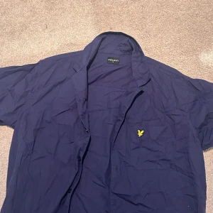 Mörkblå kortärmad skjorta Lyle & Scott - Snygg mörkblå skjorta från Lyle & Scott med korta ärmar och klassisk krage. Skjortan har knäppning framtill och en gul broderad logga på bröstet. Perfekt för dig som gillar stilrena plagg.