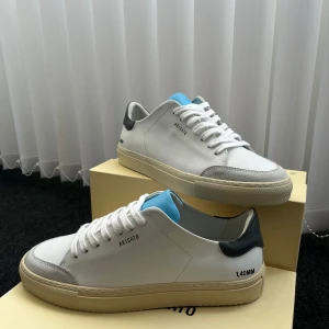 Axel Arigato vita sneakers med blå detalj - Snygga vita sneakers från Axel Arigato med grå mockadetaljer vid tån, svart häl och en ljusblå plös. Skorna har krämfärgad sula och klassisk snörning. Perfekta för dig som gillar stilrena och moderna sneakers med en twist.